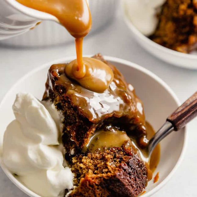 A close-up of a slice of rich chocolate cake being drizzled with thick caramel sauce, served in a white bowl with whipped cream and a wooden-handled spoon.